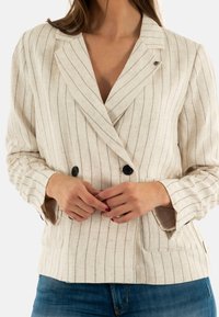 Blazer en lin rayé beige et blanc avec un design croisé, boutons noirs et manches légèrement évasées.