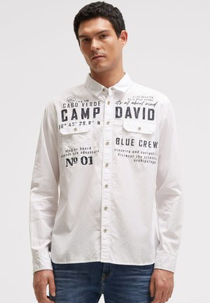 Mann trägt ein weißes Hemd mit schwarzen Textdrucken und blaue Jeans, steht vor einfachem hellem Hintergrund.