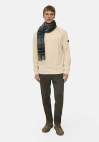 Mann mit einem cremefarbigen Pullover, einem blau-braunen karierten Schal, dunkeloliven Hosen und braunen Stiefeln, der vor einem schlichten weißen Hintergrund steht.