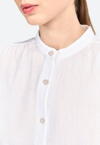 Blouse blanche texturée avec un col mandarin et des boutons décoratifs en motif coquillage. Mettez l'accent sur la légère transparence et le détail du tissu.