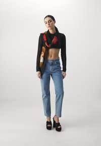 Svart netting langermet crop topp, blå høytlivsjeans og svarte platåsandaler. Skjerf med bjørneprint og røde detaljer drapert rundt halsen.