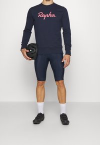 Sweatshirt navy con logo rosa "Rapha", abbinato a pantaloni ciclistici blu scuro, scarpe da ciclismo nere e un casco nero tenuto in una mano.