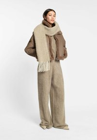 Brun vadderad jacka med shearlingdetaljer, beige oversized scarf med fransar och beiga ullbyxor i mjuk textur. Spetsiga beiga skor.