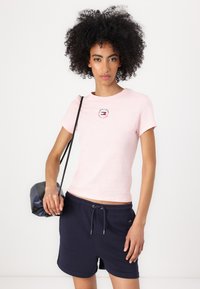 Frau mit lockigem Haar, die ein hellrosa T-Shirt mit Logo, marineblaue Shorts mit Kordelzug trägt und eine kleine schwarze Schultertasche hält.