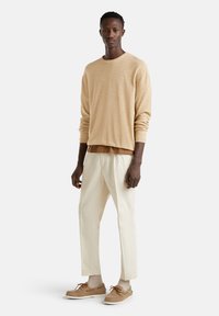 Gelber Strickpullover über einem braunen Hemd, kombiniert mit cremefarbenen Hosen und braunen Wildlederschuhen mit gestreiften Socken. Einfaches Design und Texturen.