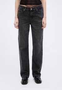 Άτομο που φοράει στενά μαύρα τζιν, μαύρα μοκασίνια και ένα κοντό μαύρο τοπ με επαναλαμβανόμενο κείμενο, σε απλό λευκό φόντο.