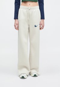 Krém színű, laza szabású melegítőnadrág, elasztikus derékkal és sötétkék hímzett logóval a bal combon.
