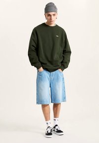 Dark green sweatshirt, light blue denim shorts, gray knit beanie, white socks with black shoes. Model standing, hands in pockets.