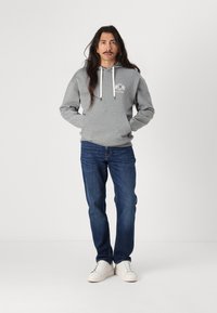 Grau Hoodie mit einem Frontlogo, Kordelzug und Kängurutasche, kombiniert mit dunkelblauen Jeans und weißen Sneakers. Glatte Stoffoberfläche.