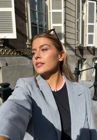 Szary marynarka w prążki na czarną koszulę, w komplecie z różową i perełkową naszyjnik. Okulary przeciwsłoneczne na włosach, miejski krajobraz z jasnymi teksturami.