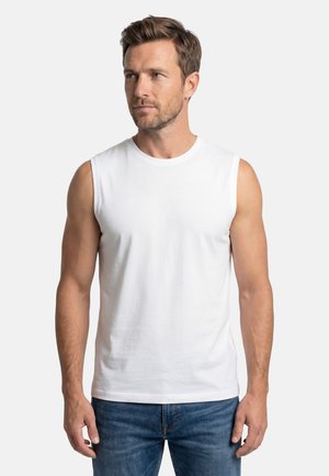 Mann mit kurzen braunen Haaren, der ein ärmelloses weißes Hemd und blaue Jeans trägt und seitlich vor einem schlichten Hintergrund schaut.