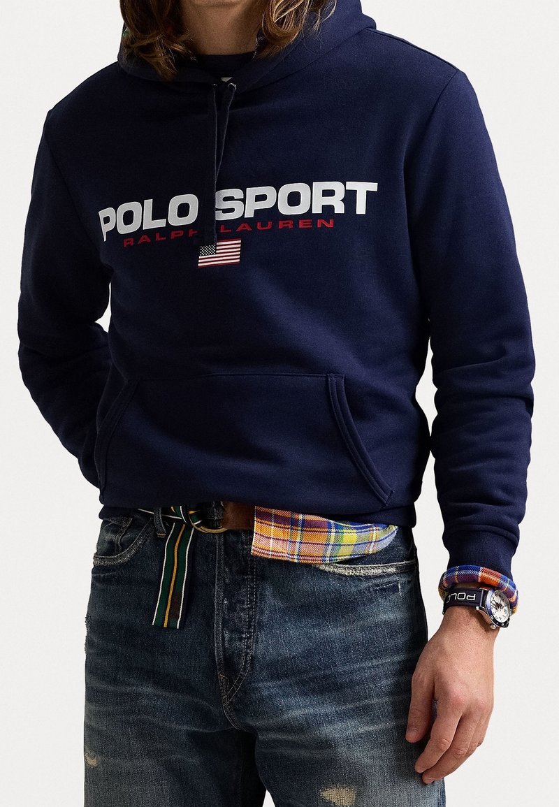 Man med marinblå Polo Sport hoodie med amerikanskt flaggmärke, rutigt skjorta instoppad i jeans, randigt bälte och en armbandsur med tygrem.