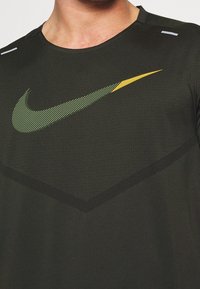 Camiseta deportiva negra de Nike con un logo en forma de chevrón en verde y amarillo, cuello redondo y tejido texturizado con acentos de costura en contraste.