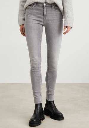 Personne portant un jean skinny gris clair, un pull en maille gris et des bottines noires épaisses, debout sur un sol uni.