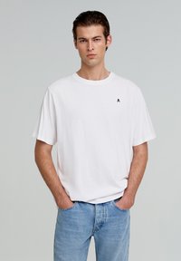 Witte katoenen t-shirt met een klassieke pasvorm, korte mouwen en een klein zwart schedel logo op de linkerborst. Gepaard met lichtblauwe jeans.