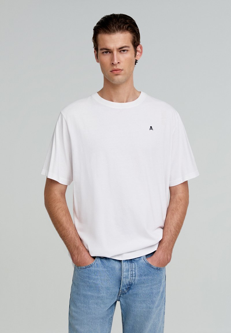 Witte katoenen t-shirt met een klassieke pasvorm, korte mouwen en een klein zwart schedel logo op de linkerborst. Gepaard met lichtblauwe jeans.