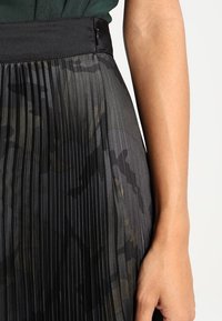 Gonna nera plissettata con una cintura in satin; presenta un sottile motivo camouflage in tonalità scure, evidenziando la texture e la profondità.