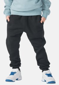 Zwarte joggingbroek met een ontspannen pasvorm, voorzien van een elastische tailleband en zijzakken. Gecombineerd met witte sportschoenen met blauwe accenten.