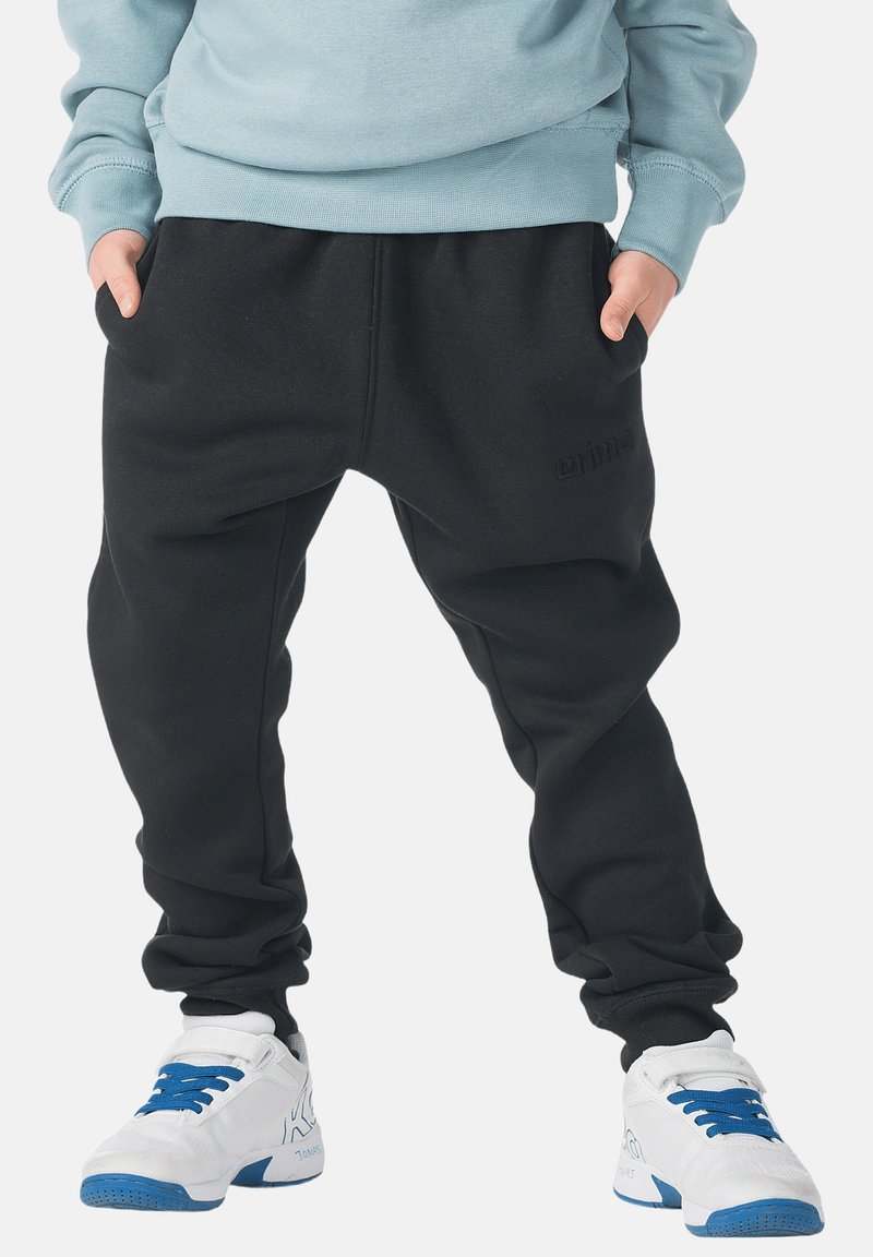 Zwarte joggingbroek met een ontspannen pasvorm, voorzien van een elastische tailleband en zijzakken. Gecombineerd met witte sportschoenen met blauwe accenten.