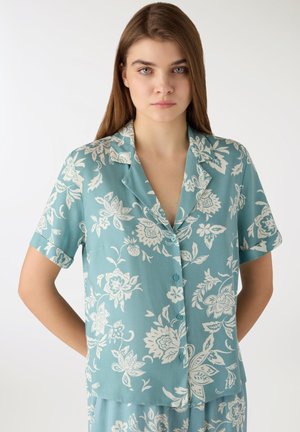 Giovane donna che indossa una camicia blu a maniche corte con bottoni e pantaloni coordinati con stampa floreale bianca, in piedi con le mani dietro la schiena.