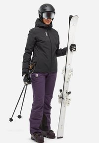 Veste de ski noire, pantalon de ski violet avec des détails en zip, gants noirs et casque de ski noir. Tient des skis blancs et des bâtons de ski avec des poignées en liège.