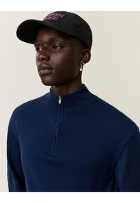 Fiatal, sötét bőrű személy, tengerészkék cipzáras pulóvert, fekete sapkát piros és kék felirattal, valamint egyetlen gyöngy fülbevalót visel.