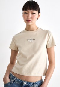 Bežinė trumpa medvilninė marškinėlių palaidinė, turinti apvalų iškirpimą ir trumpas rankoves. Priekyje yra pilka ir juoda "Calvin Klein Jeans" logotipas.