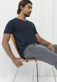 Tmavě modré tričko s krátkým rukávem a výstřihem do V vyrobené z měkkého materiálu, v kombinaci se světle šedými džíny, sedící na dřevěné  židli.