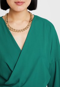 Frau trägt eine klobige goldene Kettenhalskette über einer grünen V-Ausschnitt-Wickelbluse, zeigt ihre Lippen und ihren Hals mit kurzem schwarzem Haar.