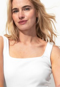 Débardeur blanc côtelé avec un décolleté carré et de larges bretelles. Le tissu est doux et légèrement texturé, offrant un ajustement près du corps au niveau des épaules.