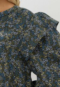 Blommig blus med en mörkgrön bakgrund prydd med blå och gula blommor, texturerad tyg och puffärmar.