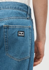 Κοντινή λήψη στην πίσω τσέπη μπλε τζιν, με ένα μικρό ασπρόμαυρο έμπλαστρο που απεικονίζει δύο σχέδια χεριών, δίπλα στο χέρι ενός ατόμου.