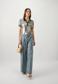 Donna che indossa una polo a maniche corte di paillettes argento, pantaloni grigi a vita alta e a gamba larga, sandali nudi e porta una borsa beige.