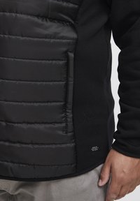 Black insulated jacket featuring a padded texture on one side and a smooth fabric on the other, with a small label near the hem.