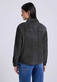 Vrouw in een donkergrijze corduroy jas en een blauwe spijkerbroek, met haar naar achter gekeerd, steil donker haar en grote zilveren hoopoorbellen.