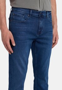 Jeans in denim blu scuro con una vestibilità slim, dotati di cinque tasche, accessori in metallo e una finitura leggermente texturizzata.