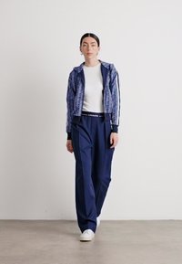 Giacca a zip navy con motivo astratto azzurro, abbinata a pantaloni larghi navy e sneaker bianchi. Tessuto morbido, design sportivo.