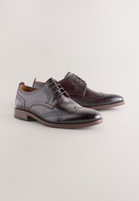 Chaussures habillées en cuir marron avec détails brogues, design plat à lacets, bout pointu, et talon moyen. Texture lisse avec accents perforés.