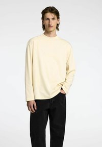 Majica s dolgimi rokavi v bledo rumeni barvi iz mehkega bombažnega materiala z sproščenim krojem. V kombinaciji z črnimi kavbojkami, kar ponuja preprost in sproščen videz.