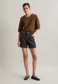 Bruin getextureerde gebreide trui met korte mouwen, gecombineerd met hoge zwarte denim shorts. Het model draagt zwarte platte schoenen en een zilveren ketting.