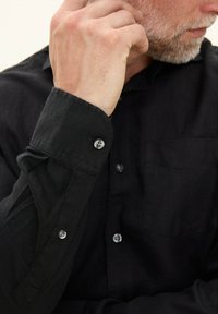 Zwarte linnen overhemd met een button-down kraag, een enkele borstzak en knoopmanchetten met transparante kunststof knopen. Textuurdetail van de stof zichtbaar.