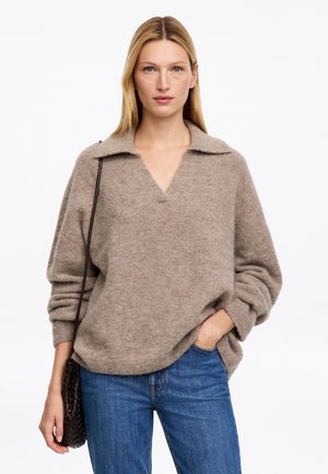 Femme aux cheveux blonds et raides portant un pull ample taupe, un jean bleu et portant un sac à bandoulière tissé foncé, debout devant un fond blanc.