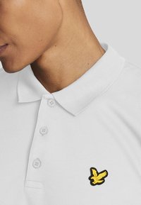 Polo shirt bianco con tessuto testurizzato, caratterizzato da un logo di uccello giallo sul lato sinistro del petto, doppio colletto con bottoni e design del colletto classico.