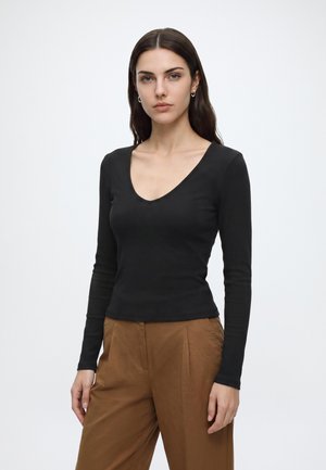 Femme aux longs cheveux foncés portant un haut noir à manches longues avec un col en V et un pantalon marron, debout devant un fond clair uni.