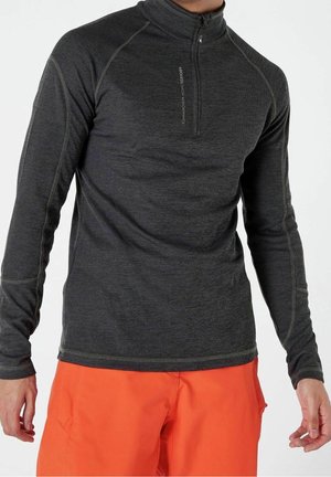 Homme portant un haut gris foncé à manches longues avec fermeture éclair quart, et un pantalon orange vif, debout avec les bras détendus le long du corps.