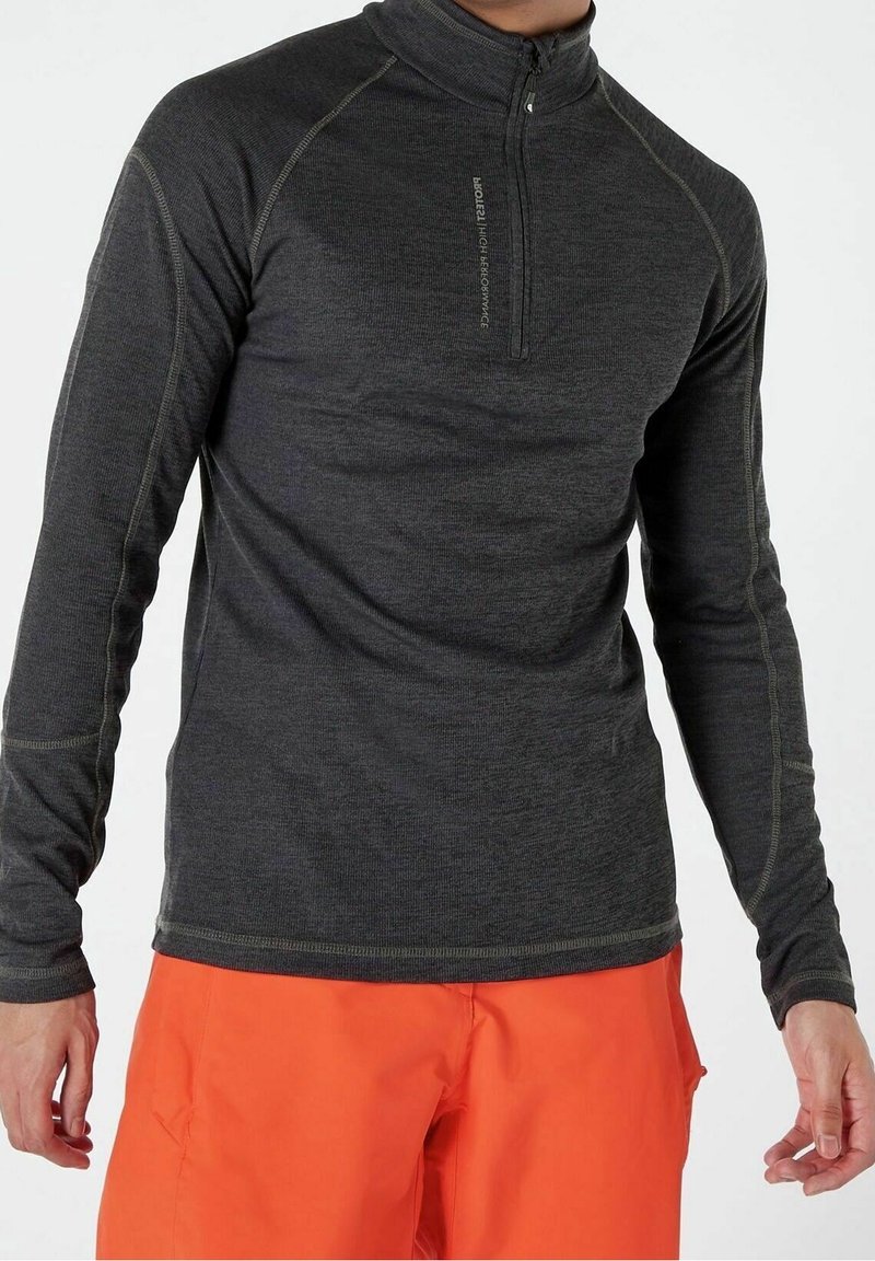 Homme portant un haut gris foncé à manches longues avec fermeture éclair quart, et un pantalon orange vif, debout avec les bras détendus le long du corps.
