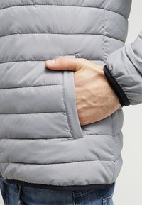 Mano inserita nella tasca laterale di una giacca trapuntata grigia chiara indossata con jeans blu.