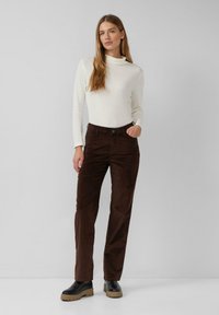 Pantaloni in velluto a coste marroni con vestibilità dritta, abbinati a una maglietta a maniche lunghe color crema. L'outfit presenta un colletto alto e una leggera texture.