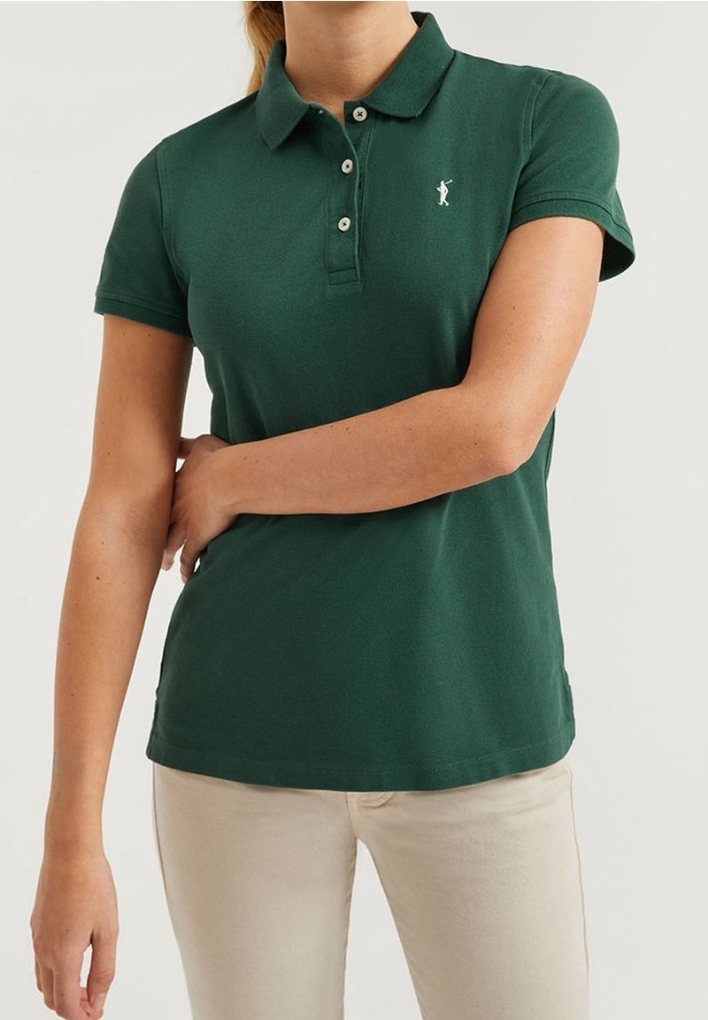Femme portant un polo à manches courtes vert foncé avec un pantalon beige, un bras croisé sur la taille, fond clair uni.