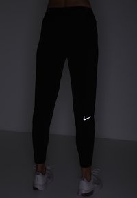 Černé sportovní kalhoty s přiléhavým střihem, vystupující s reflektující bílou Nike logem na levé noze a hladkým, elastickým textilním povrchem.